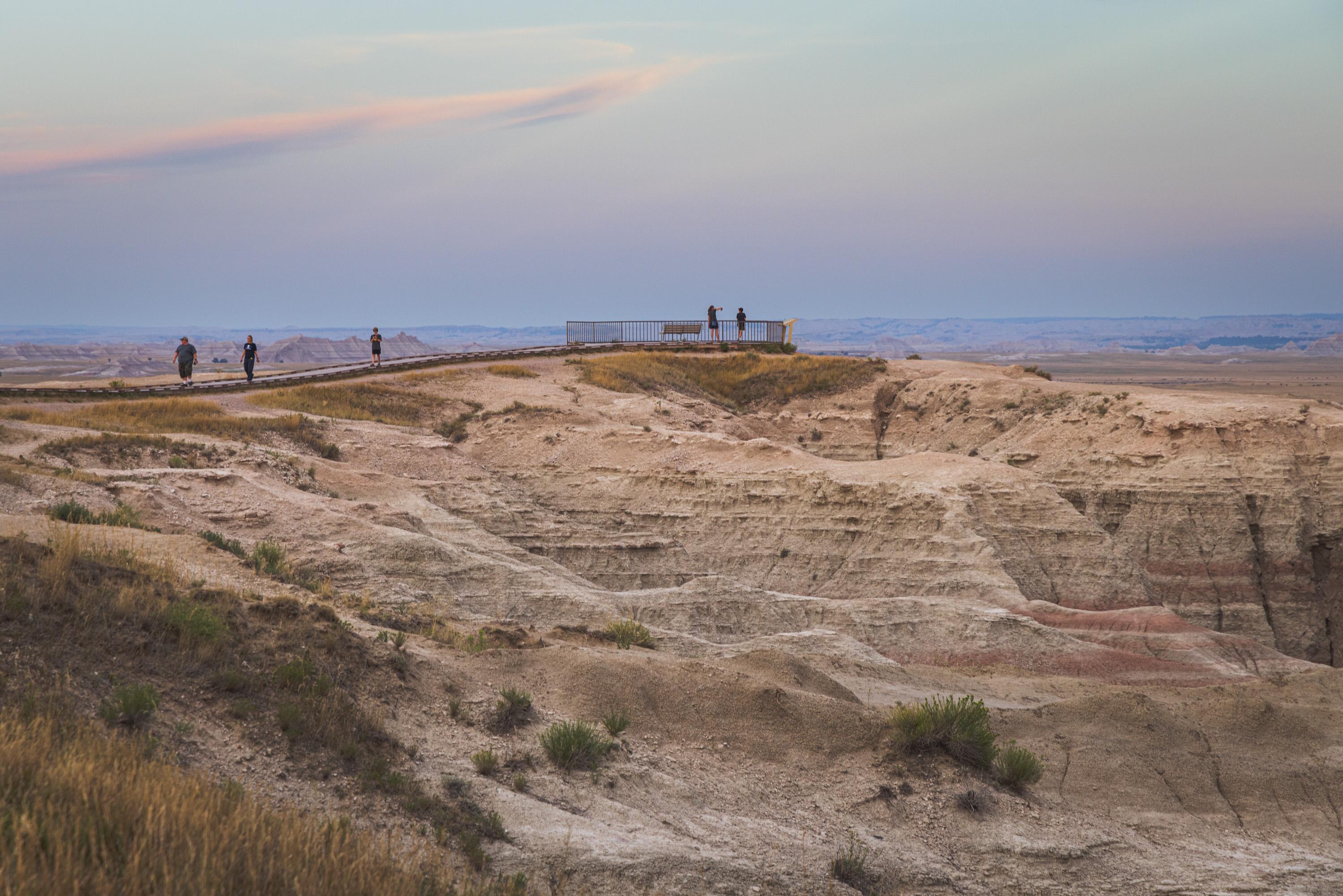 People take in a view in an observation area atop a rocky hill as others walk back on the path leading to the observation area