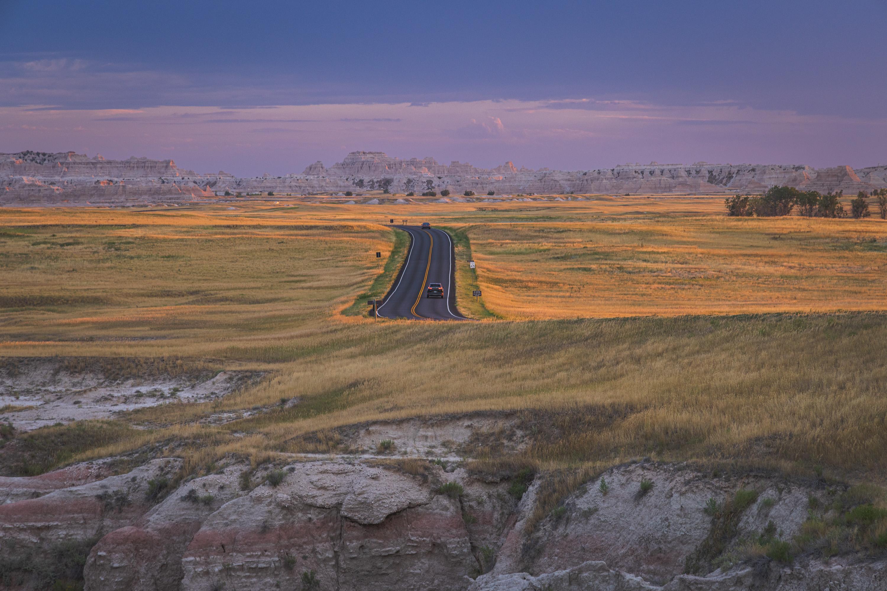The main road in the park takes a turn, giving the viewer a sightline directly down the road as a single car drives into the park at sundown. The grasslands on either side of the road are lit with diffuse light, and the sky behind above rocky hills is a subtle purple-blue