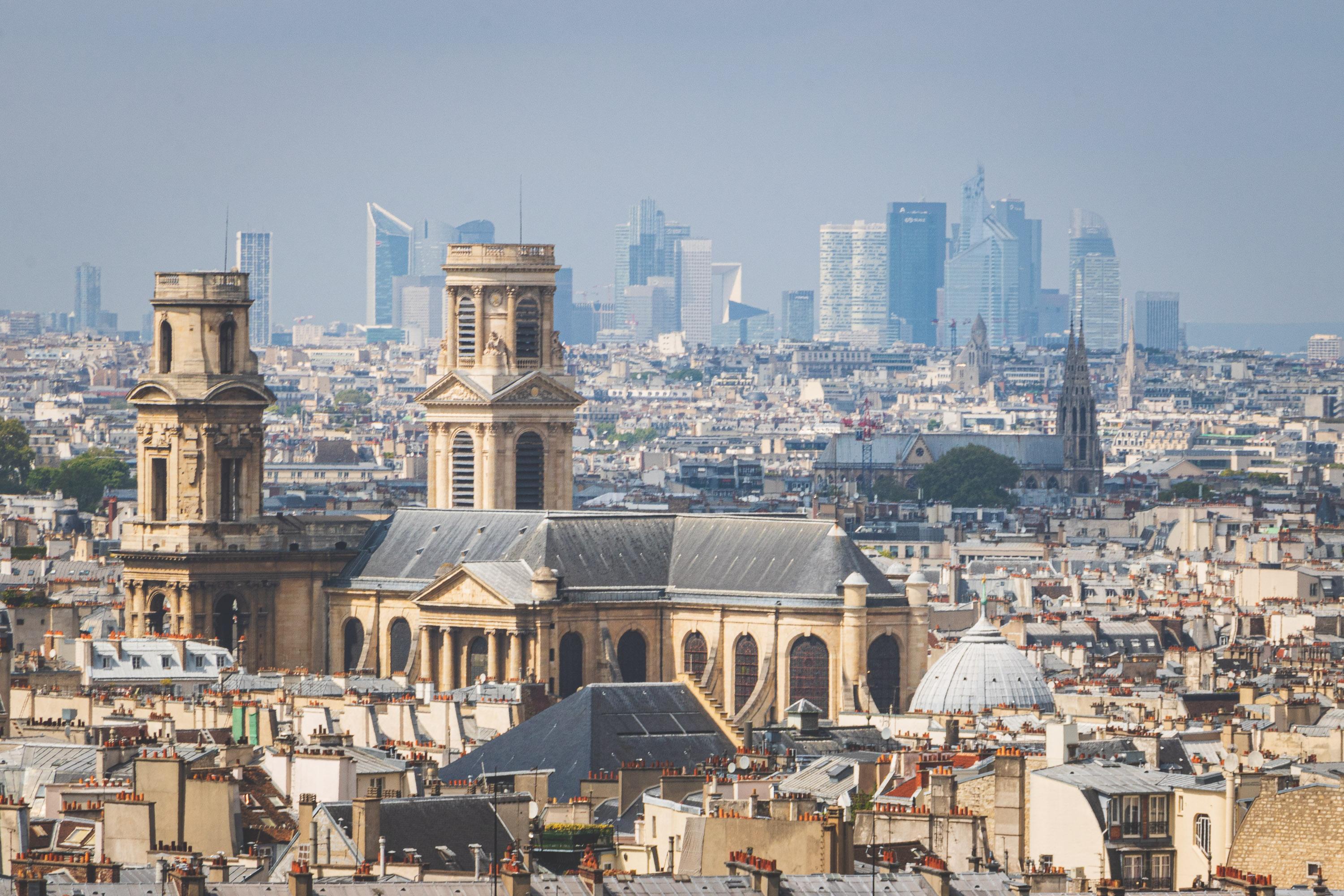 A view of an old, gothic-style church in a neighborhood, set against the backdrop of La Defense, Paris' modern business district filled with glass and steel skyscrapers