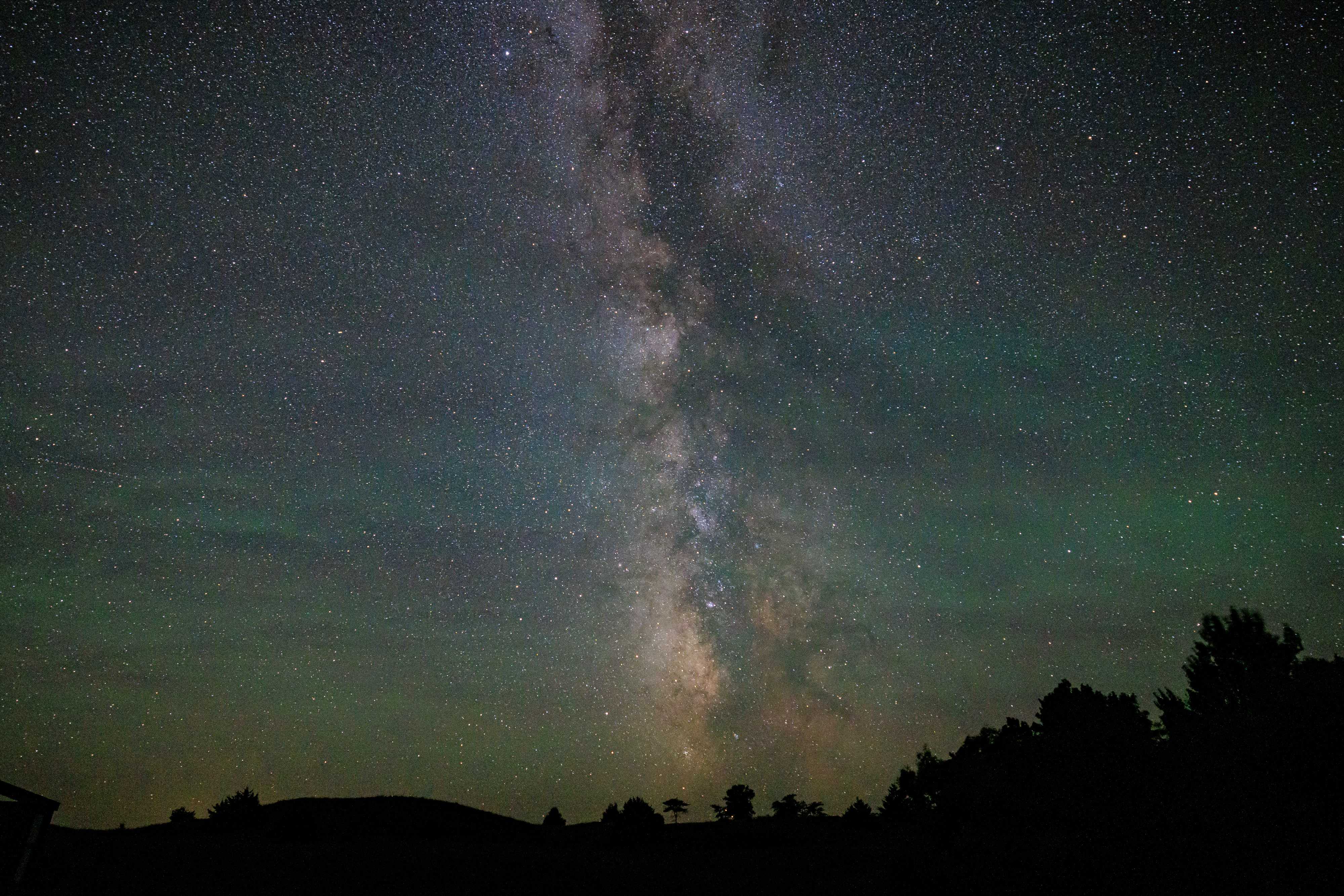 The Milky Way is visible rising from the tree-lined horizon into the dark of the night sky, a clear band of almost infinite stars dense in the sky. A faint green sheen overlays the photo, which is a unique phenomenon called skyglow, visible after dark in only the least light polluted skies on earth.