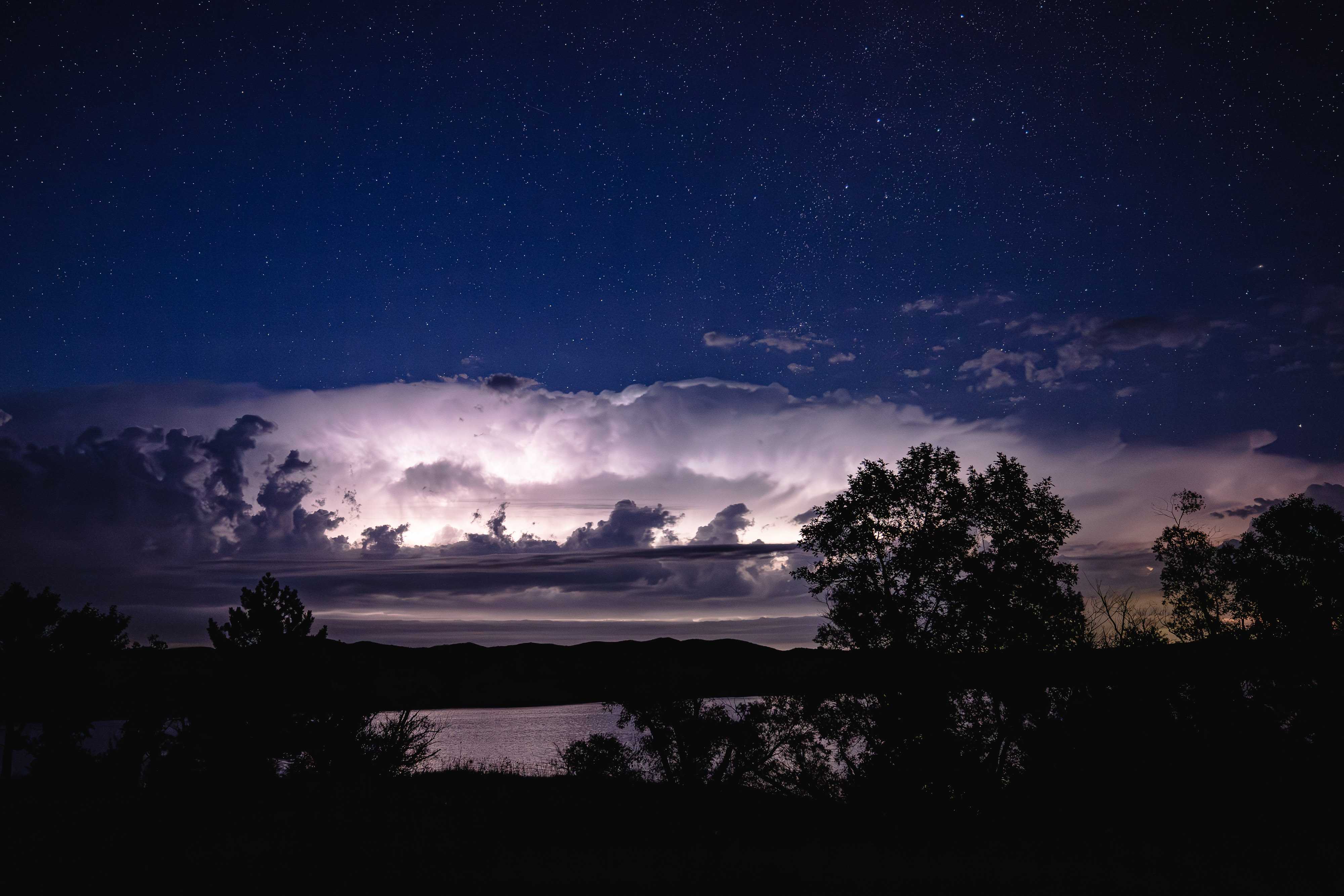 Fluffy clouds are lit by lightning flashes in front of a deep blue sky filled with stars