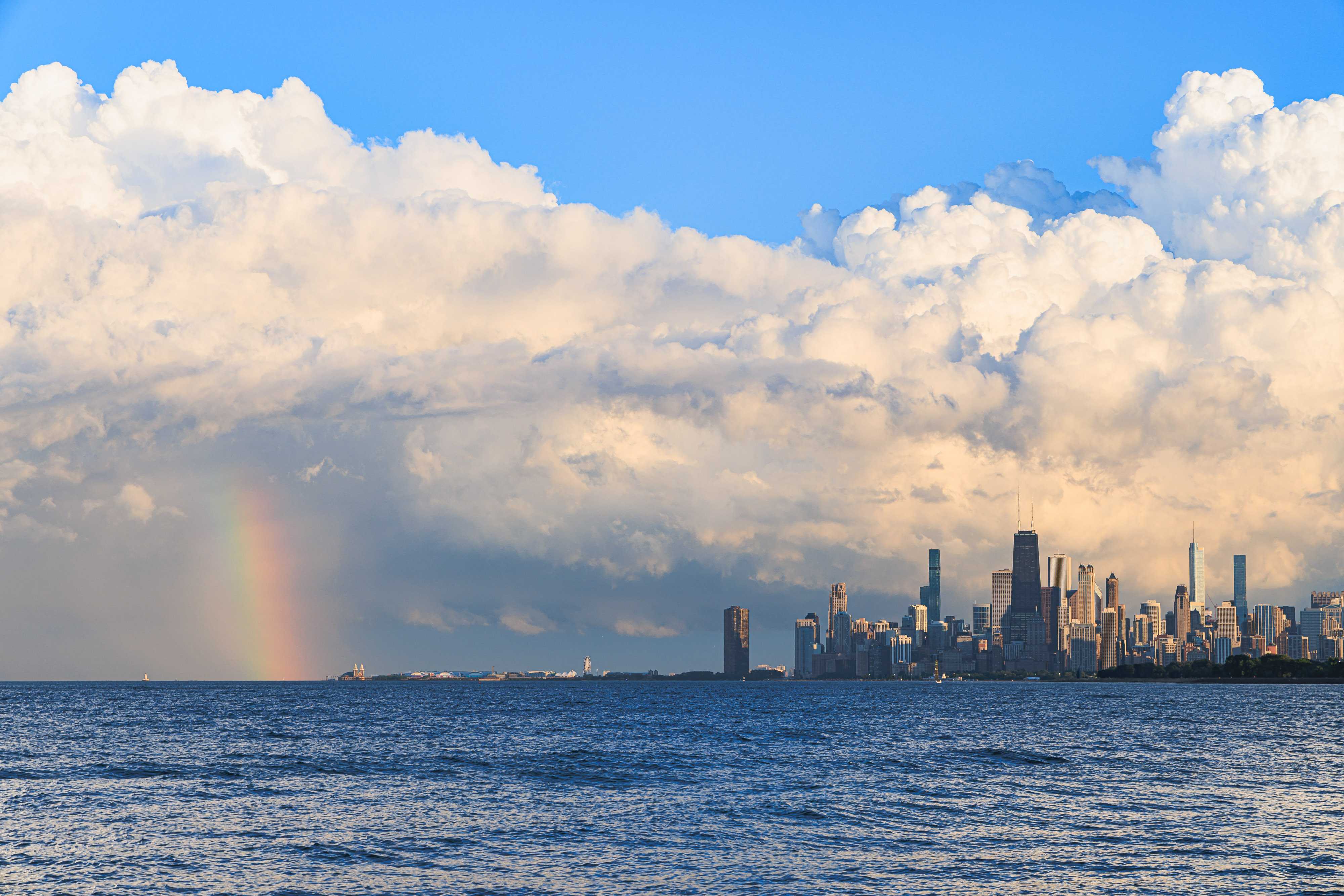 Elaborately twisted storm clouds tower the city skyline, which is set to the right of the image seemingly rising from the choppy waters of Lake Michigan. The city is lit brightly, as the dark clouds of a storm blow away to reveal a sunny day. To the left of the image, a bright rainbow shines.