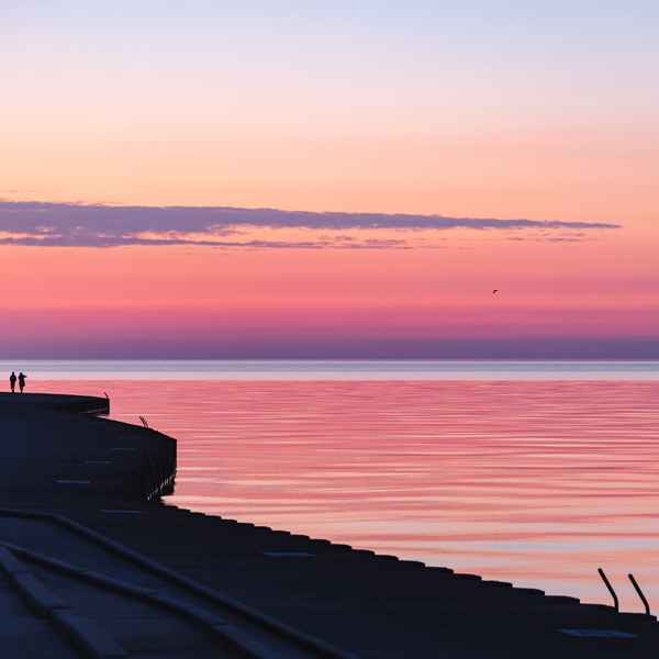 The sky is filled with orange and pink tones, blending smoothly into the gentle waves of Lake Michigan where water meets sky. To the left of the image, the silouhette of a couple walking into the sunrise along the sharp lines of the concrete revetments that line the lakeshore. 